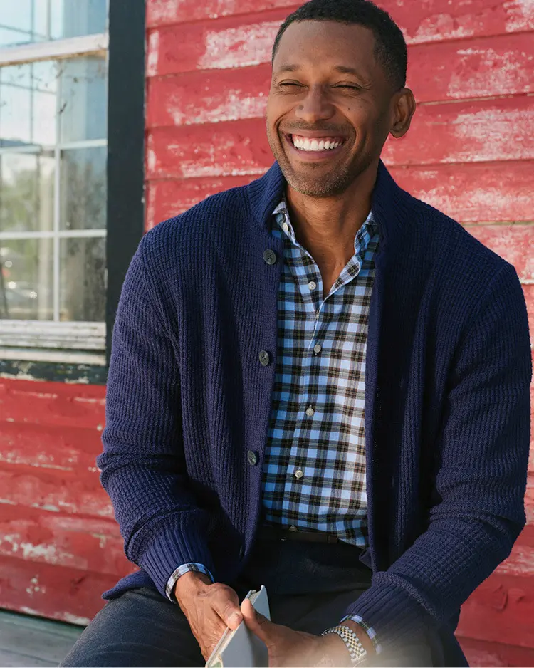 man in a plaid shirt and sweater in front of a house